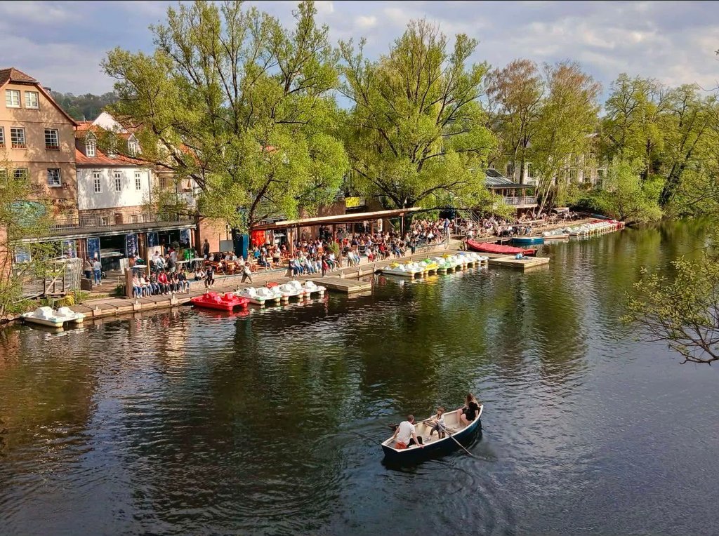 Blick über die Lahn auf das belebte Ufercafé mit voll besetzter Terrasse, bunten Tretbooten am Steg und einem Ruderboot auf dem Wasser