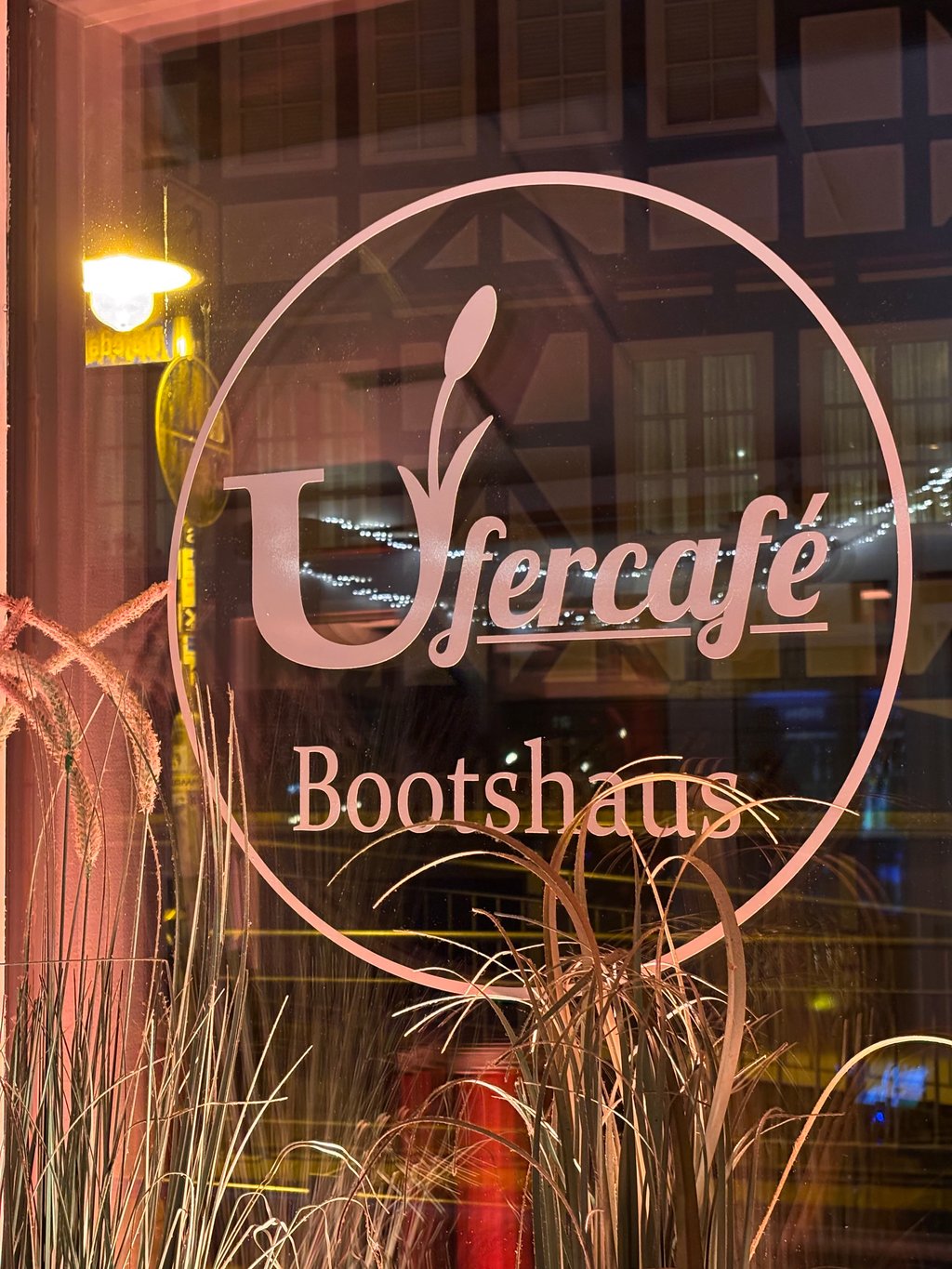 Rundes Ufercafé-Logo auf dem Fenster mit Pampasgras und Spiegelung der Marburger Fachwerkhäuser